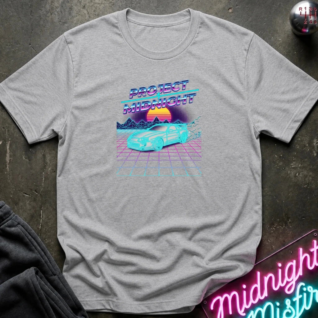 A light gray heather graphic t-shirt laid flat on a dark textured surface. The shirt features a retro 80s synthwave graphic with the text 'PROJECT MIDNIGHT' in neon purple and blue, above a cyan car on a pink grid with a sunset.