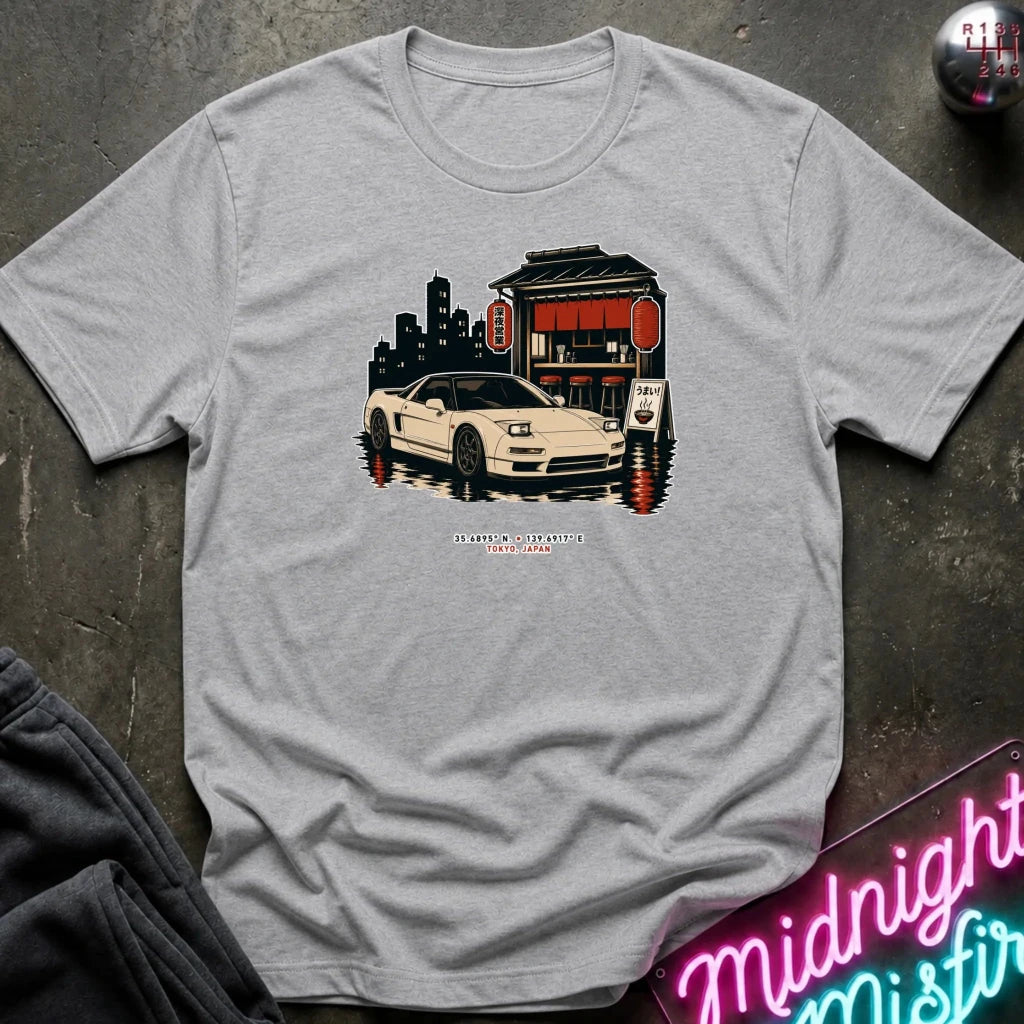 A light gray crewneck t-shirt laid flat on a dark textured surface, featuring a graphic print of a white sports car parked in front of a red-roofed building with a city skyline in the background. 
