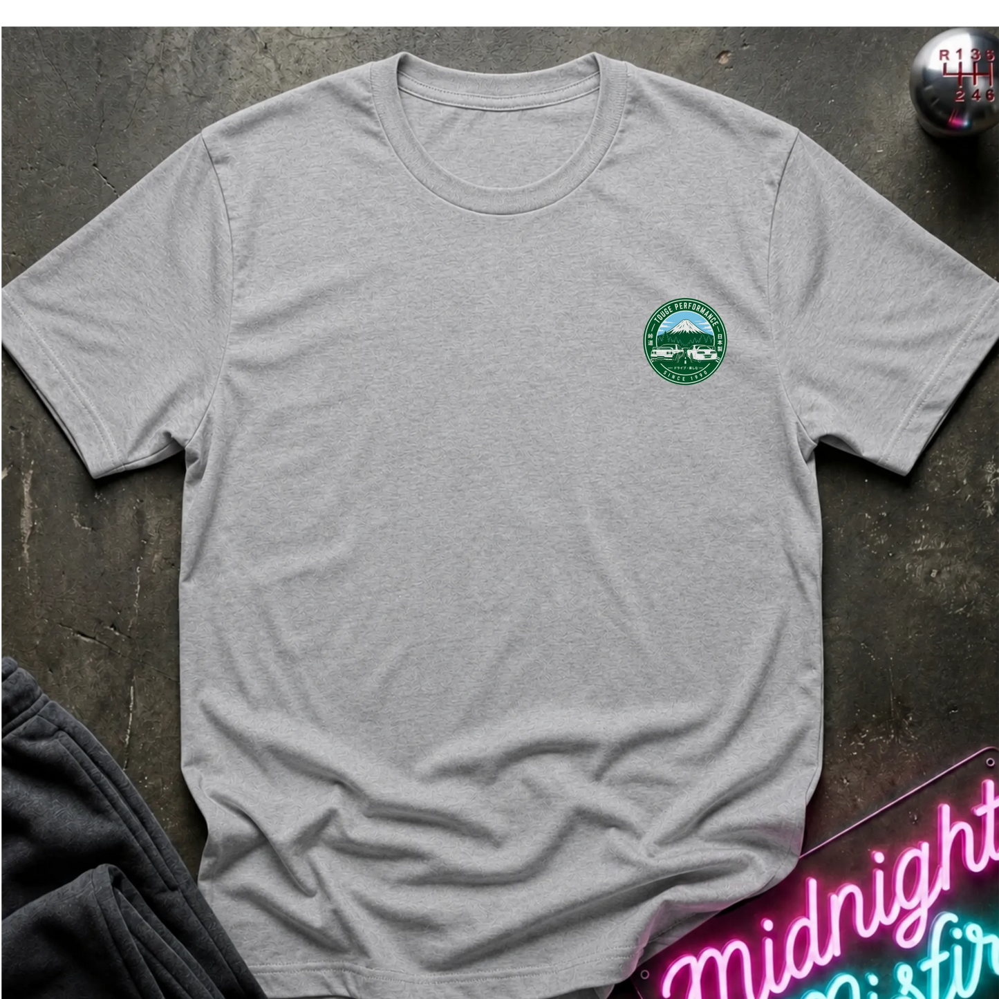 Gray short-sleeve graphic T-shirt laid flat on a dark textured surface. The shirt features a small circular green and white print logo  with a mountain, two cars, and the text 'TOUGE PERFORMANCE' and 'SINCE 1990'.