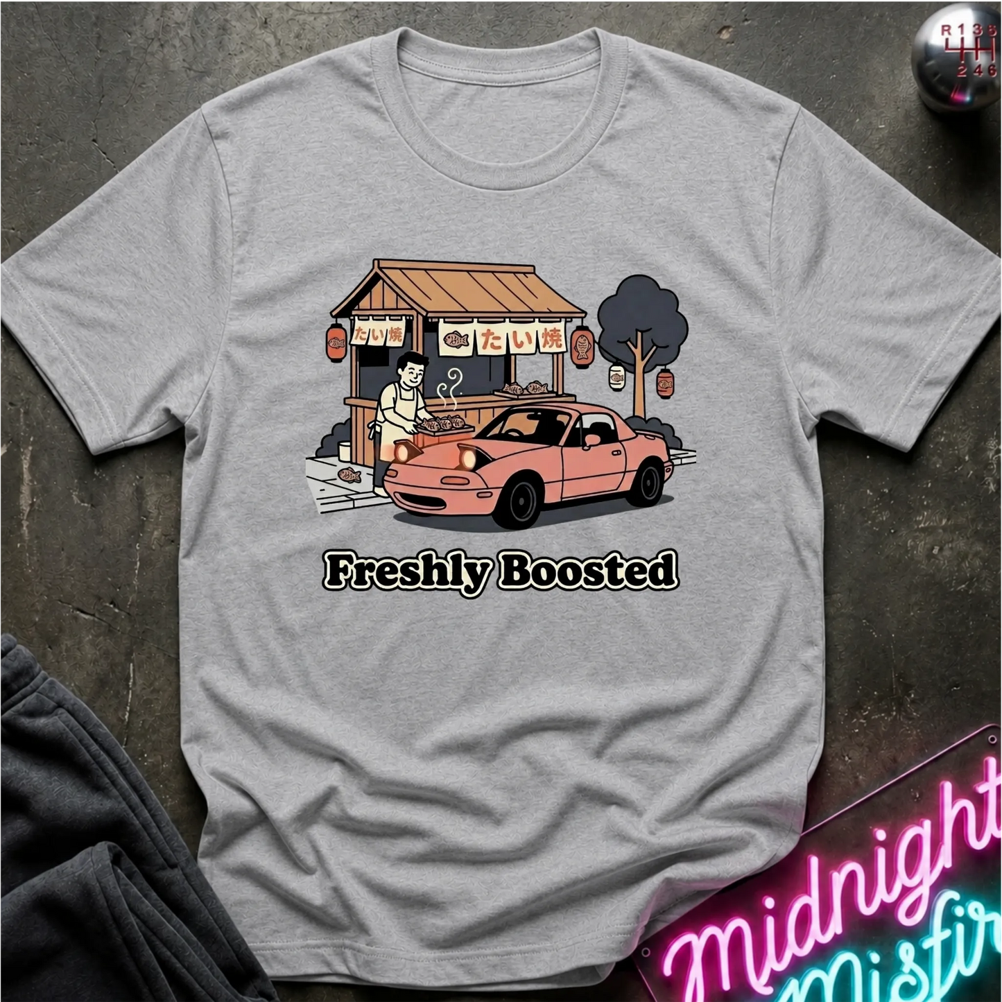A light gray heather graphic t-shirt laid flat on a dark textured surface. The graphic features a pink convertible car parked in front of a street food stall  and the text 'Freshly Boosted' printed below.