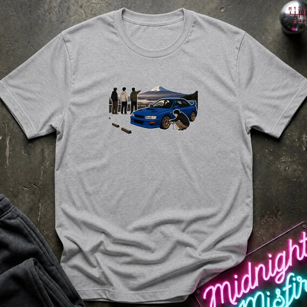 A light heather gray crewneck t-shirt laid flat on a dark textured surface. The shirt features a graphic print of a blue sports car with a driver and spectators in front of a mountain landscape.