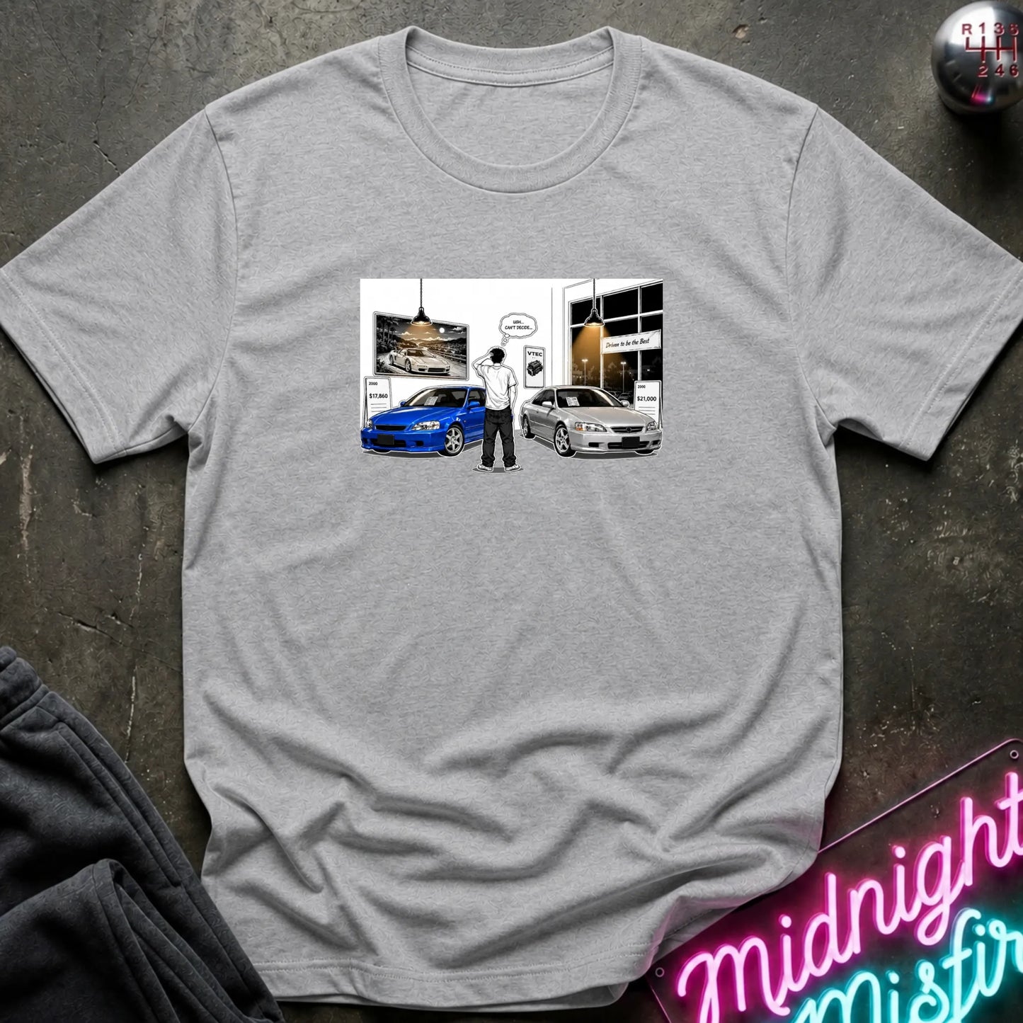 A heather gray short-sleeve t-shirt laid flat on a dark textured surface, featuring a rectangular graphic print on the chest with a cartoon figure standing between a blue car and a silver car.