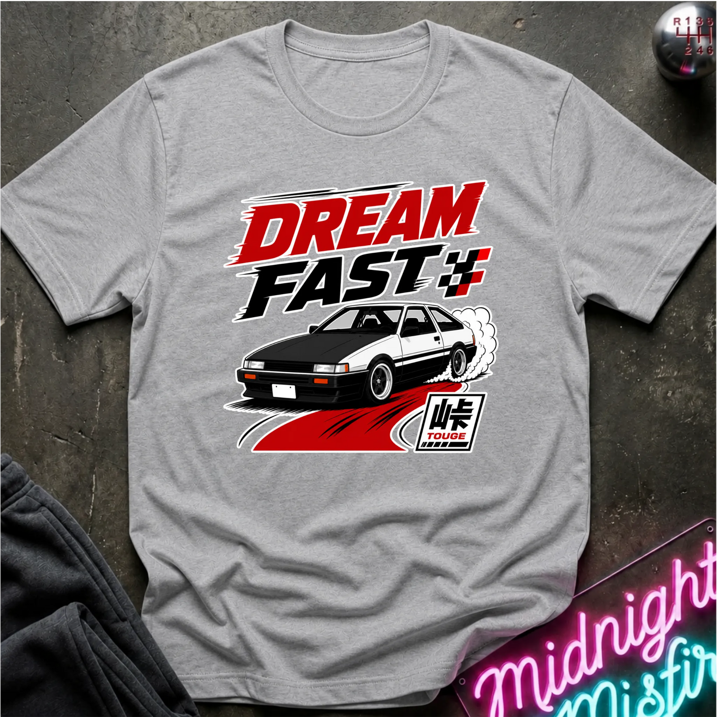 A light gray crewneck t-shirt featuring a 'Dream Fast 4' graphic print with a black and white sports car, red and white text, laid flat on a dark textured surface.