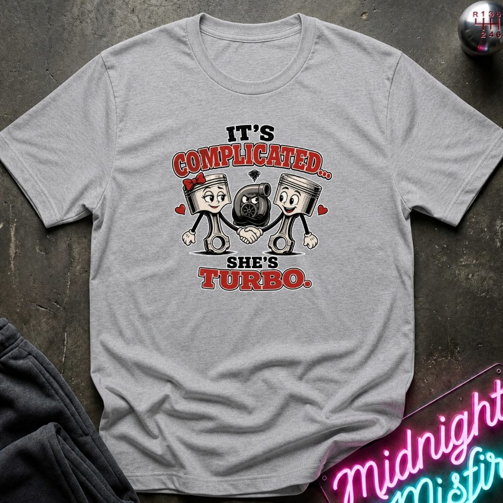 A light gray graphic t-shirt laid flat on a dark textured surface. The shirt features a humorous graphic with two cartoon pistons shaking hands, a black turbo in the middle, and the text 'IT'S COMPLICATED.