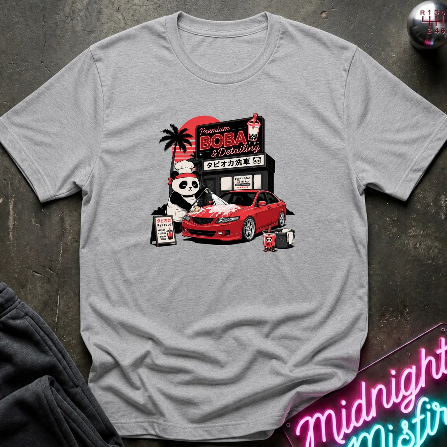 A light gray crewneck t-shirt featuring a colorful graphic print of a panda working at a red car wash shop next to a red sports car, laid flat on a dark textured surface.