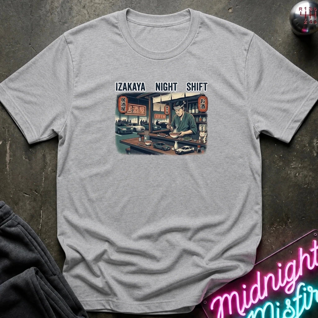 A light gray heather crewneck t-shirt featuring a vintage-style graphic of an izakaya night shift worker with the text 'IZAKAYA NIGHT SHIFT' above it. The shirt is styled flat on a dark textured surface with a neon sign and dark pants partially visible.