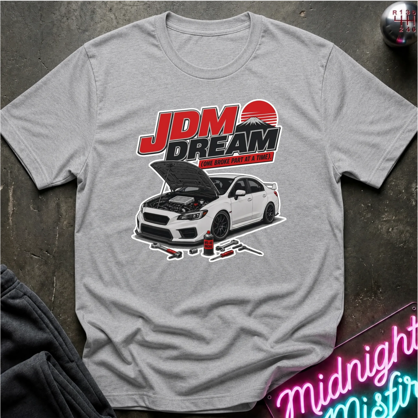 A light gray heather crewneck t-shirt featuring a graphic print of a white sports car with its hood open, under the text 'JDM DREAM ENGINEERS'. The shirt is laid flat on a dark textured surface with a neon sign and clothing partially visible in the corners.