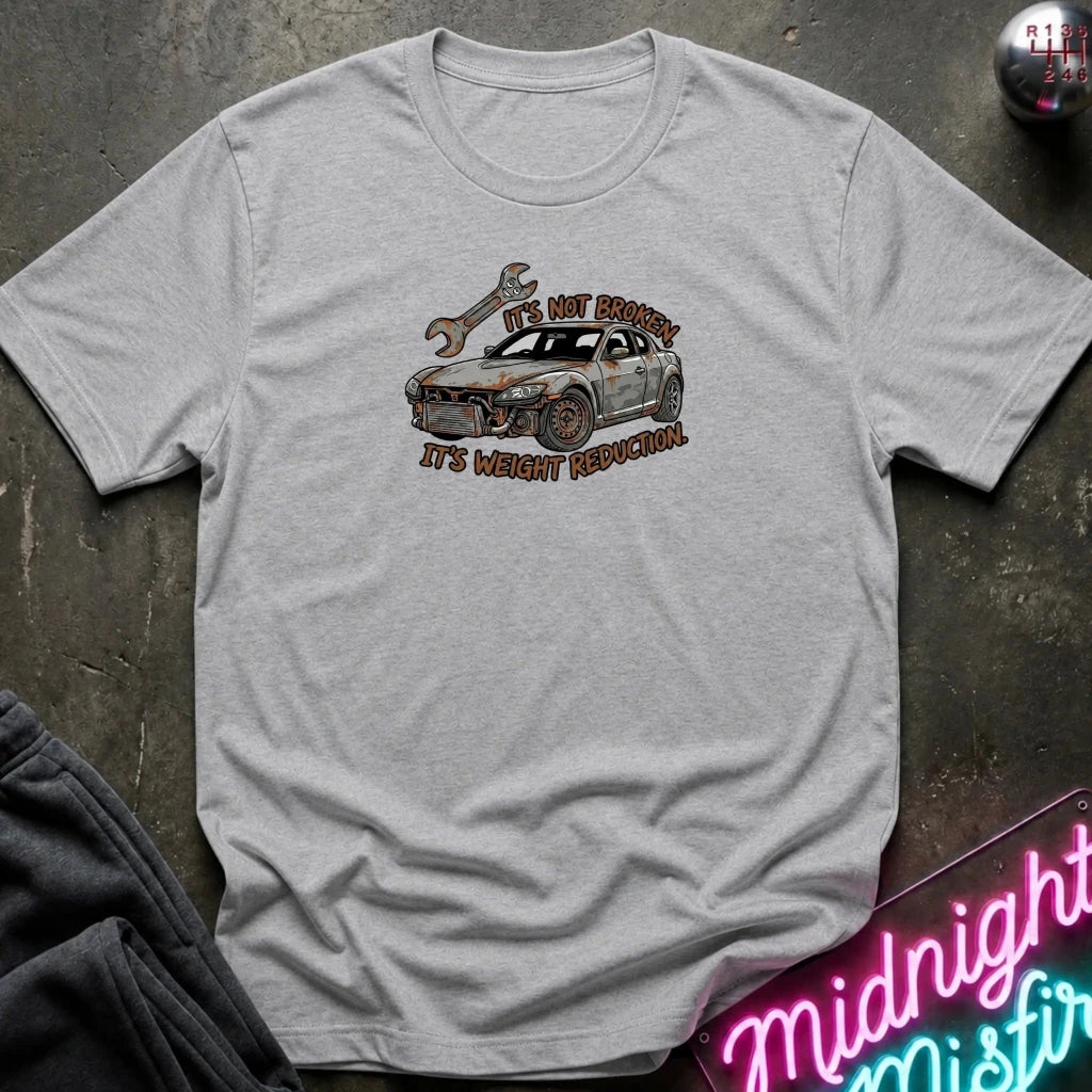 A light heather gray crewneck t-shirt laid flat on a dark textured surface. The shirt features a graphic of a white sports car with a rusted front bumper, an oil drop, and a wrench, with the text 'IT'S NOT BROKEN