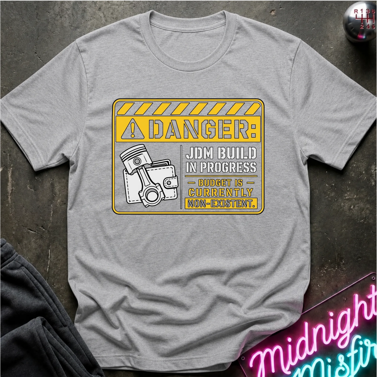A light gray short-sleeve graphic T-shirt laid flat on a dark textured surface. The shirt features a yellow and white warning-sign style print with the text 'DANGER: JDM BUILD IN PROGRESS - BUDGET IS - CURRENTLY NON-EXISTENT'.