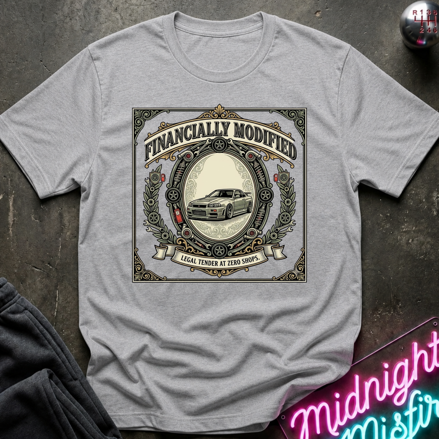 A light gray crewneck t-shirt with a vintage-style graphic print reading 'FINANCALLY MODIFIED' and 'LOCAL TENDER AT ZERO SHOPS' around an illustration of a classic car. The shirt is displayed in a flat-lay style on a dark textured surface with a neon sign and dark pants partially visible.