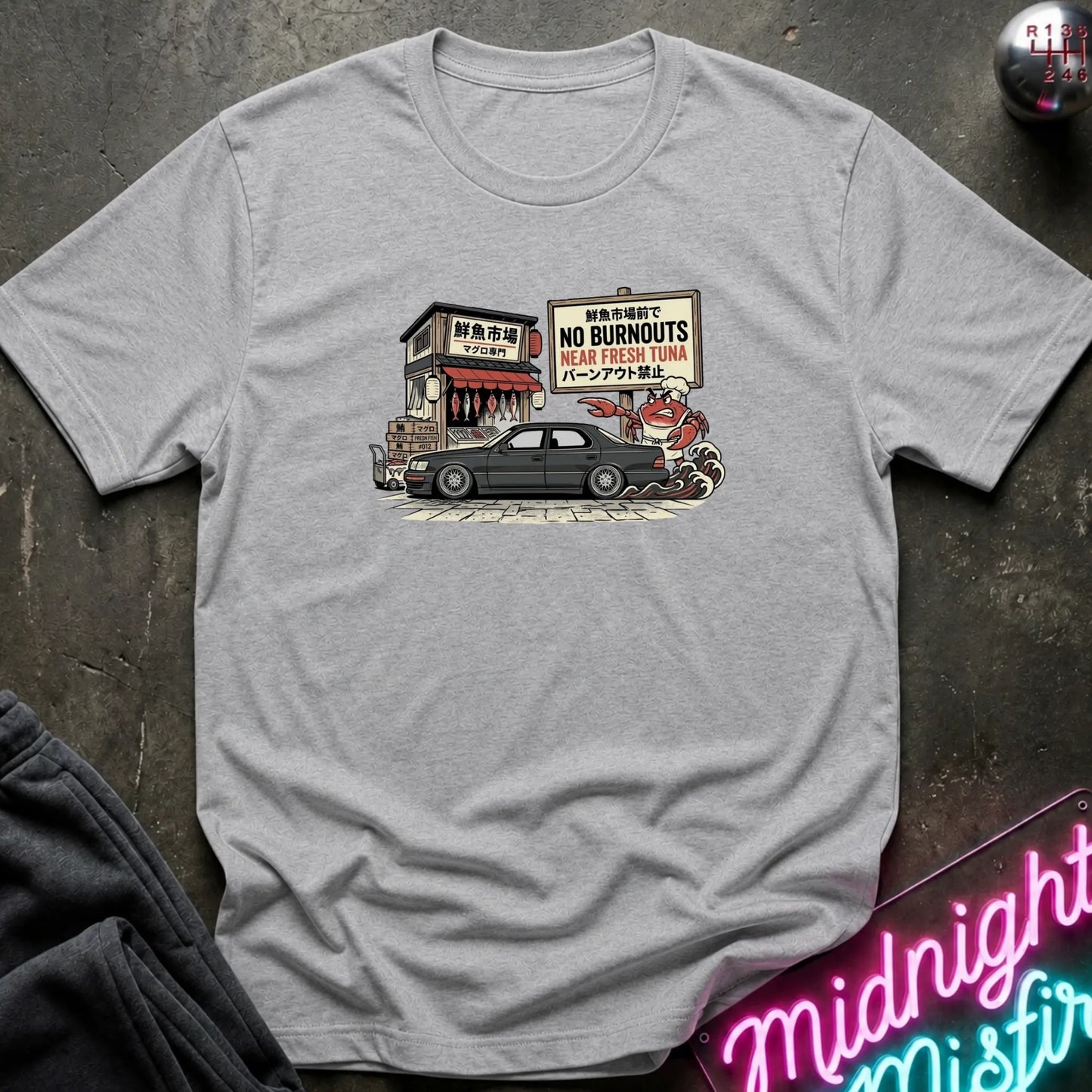 A light gray crewneck t-shirt featuring a vintage-style graphic print of a black sedan parked in front of a storefront with a 'No Burnouts' sign, laid flat on a dark textured surface with decorative props.