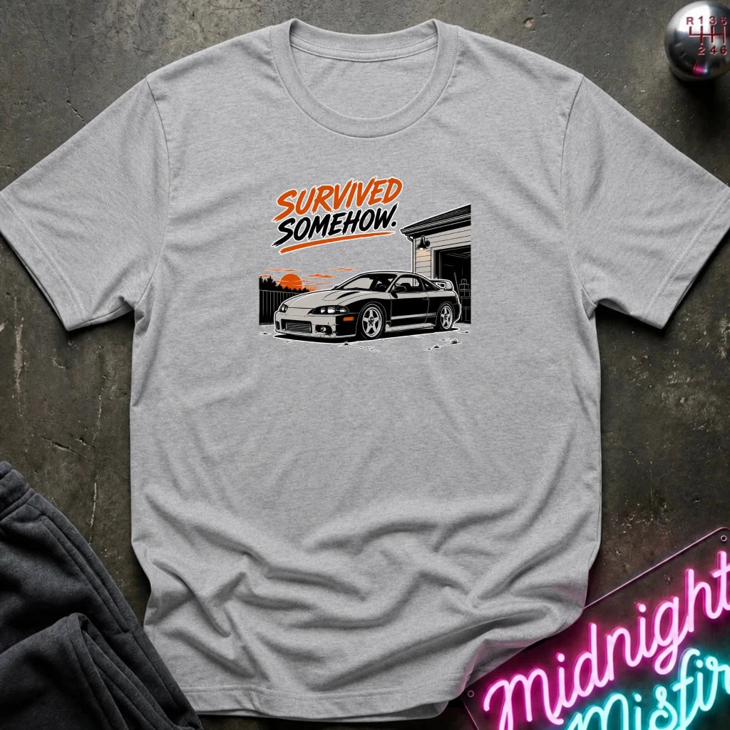 A light heather gray crewneck t-shirt laid flat on a dark textured surface. The shirt features a graphic print of a black sports car parked in front of a house with the text 'SURVIVED SOMEHOW' in orange and black.