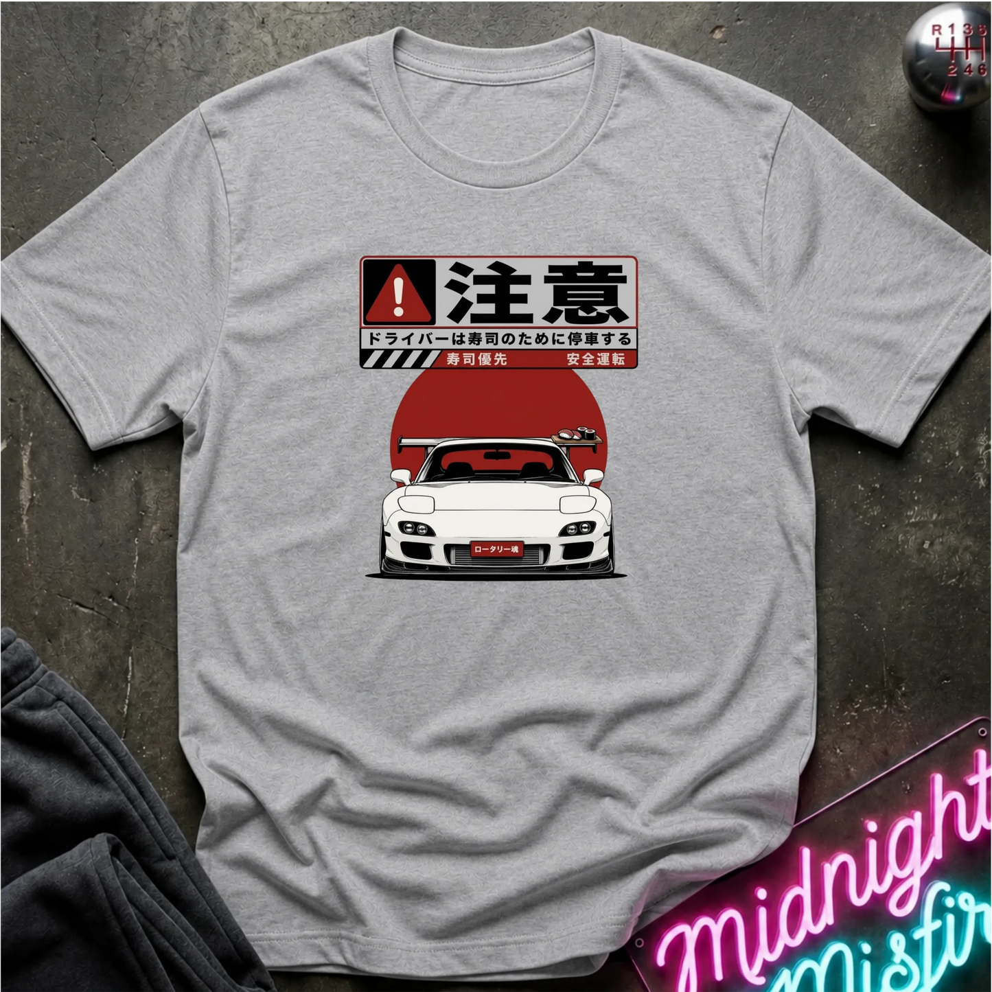 A light gray heather crewneck t-shirt featuring a graphic print of a white sports car in front of a red circular background with Japanese text. The shirt is laid flat on a dark textured surface with a neon sign and clothing prop partially visible.