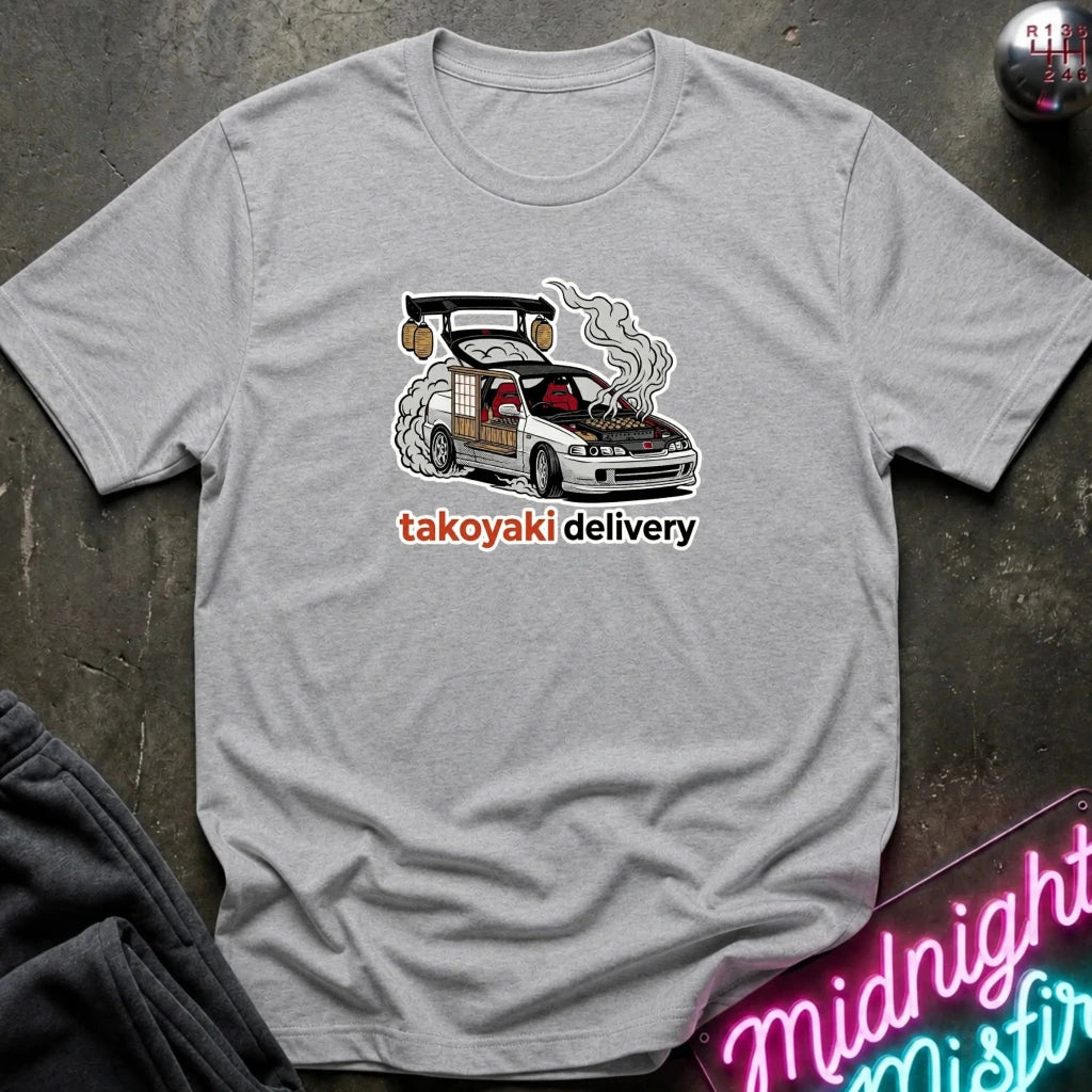 A light gray crewneck t-shirt laid flat on a dark textured surface, featuring a graphic of a smoking white sports car with the text 'takoyaki delivery' printed below it.