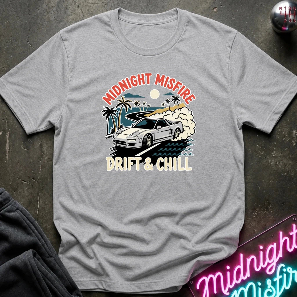 A heather gray short-sleeve t-shirt with a retro-style graphic of a white sports car drifting on a beach with palm trees, under a sunset. The graphic text reads 'MIDNIGHT MISFIRE' and 'DRIFT & CHILL'.
