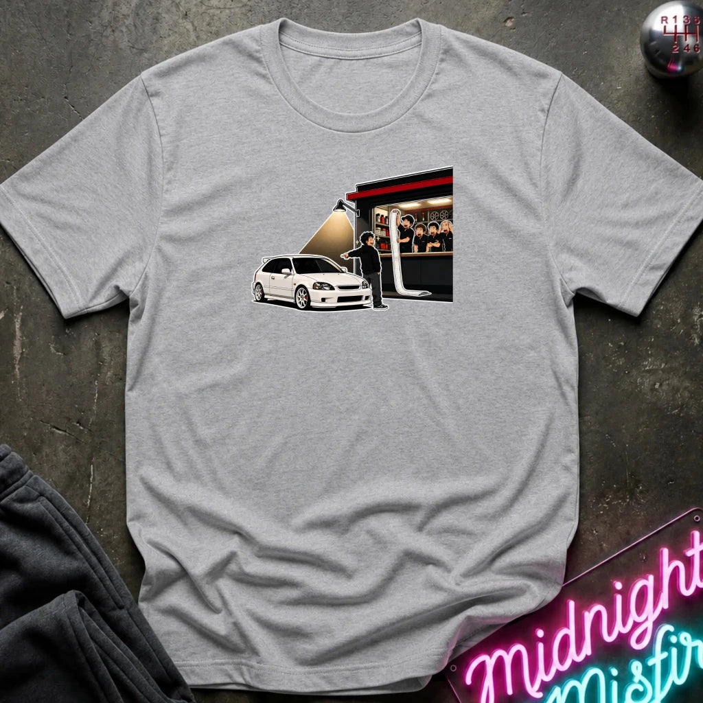 A heather gray crewneck t-shirt laid flat on a dark textured surface, featuring a graphic print of a white car in front of a neon-lit shop