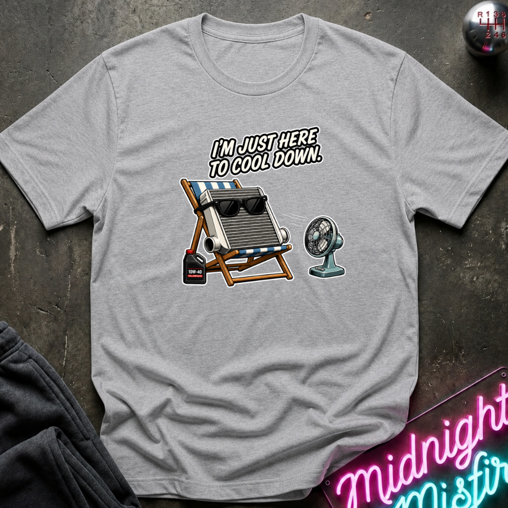 A light gray short-sleeve t-shirt laid flat on a dark textured surface, featuring a graphic of a folding beach chair with a cooler and a small fan, with the text 'I'M JUST HERE TO COOL DOWN.' A neon sign and folded dark fabric are partially visible around the edges.