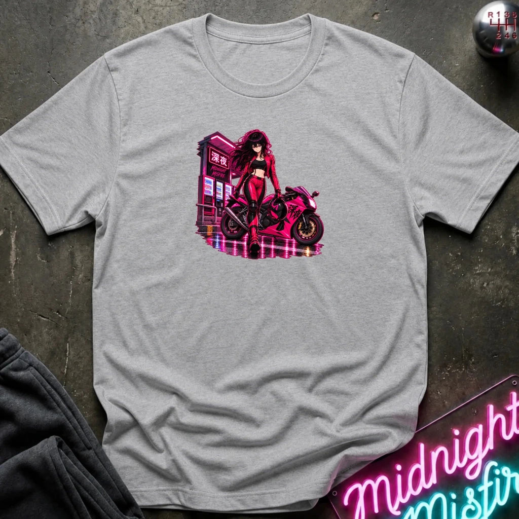 A light heather gray short-sleeve t-shirt laid flat on a dark textured surface, featuring a colorful graphic print of a woman in a red outfit standing next to a pink motorcycle with neon-style building elements. 
