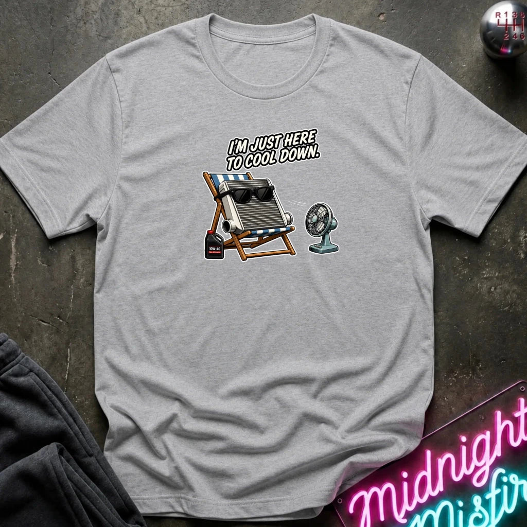 A light gray crewneck t-shirt laid flat on a dark textured surface. The shirt features a graphic of a beach chair with sunglasses, a small fan, and the text 'I'M JUST HERE TO COOL DOWN!'.