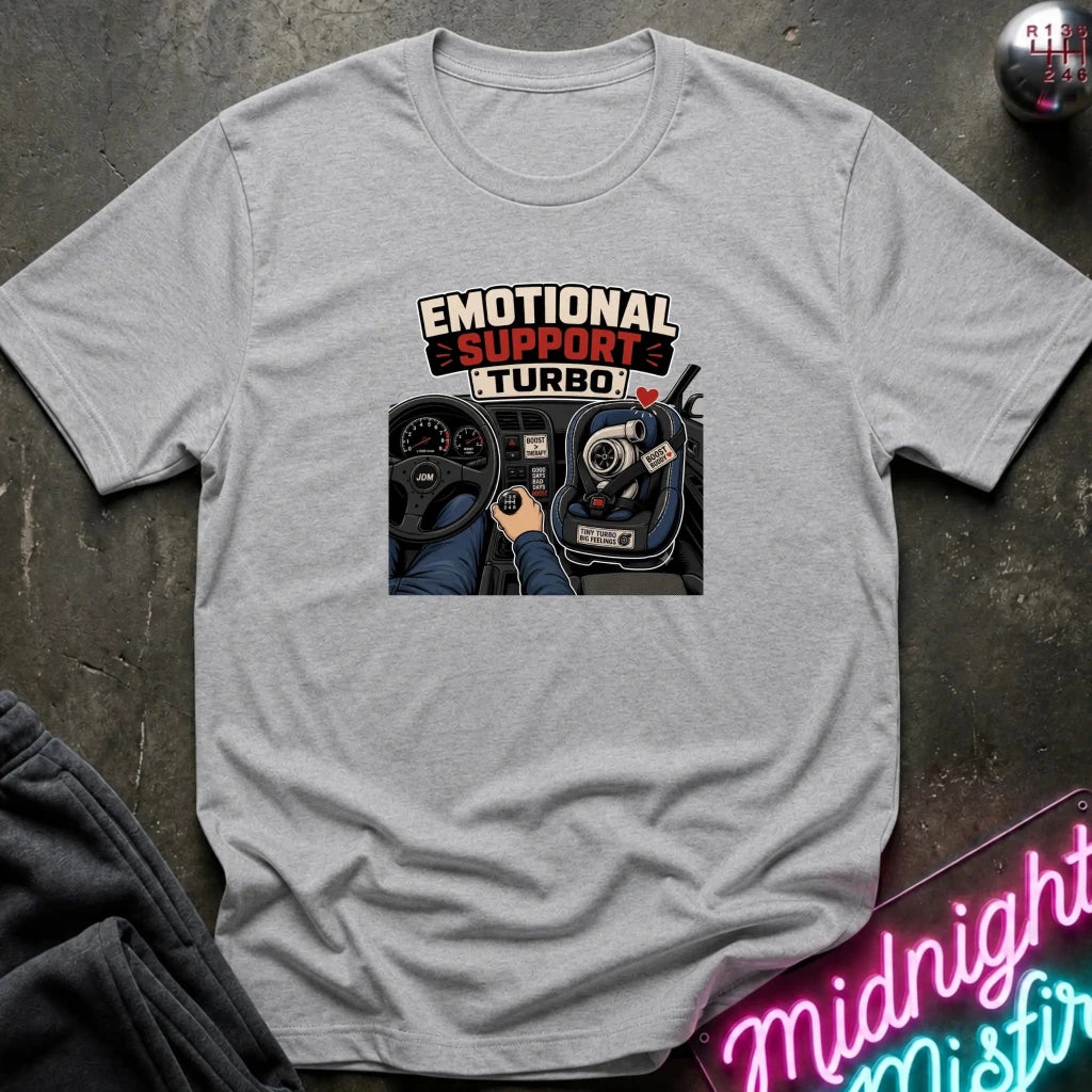 A light gray crewneck t-shirt laid flat on a dark textured surface, featuring a graphic print of a car dashboard with the text 'EMOTIONAL SUPPORT TURBO!' and a steering wheel and gear shift illustration.
