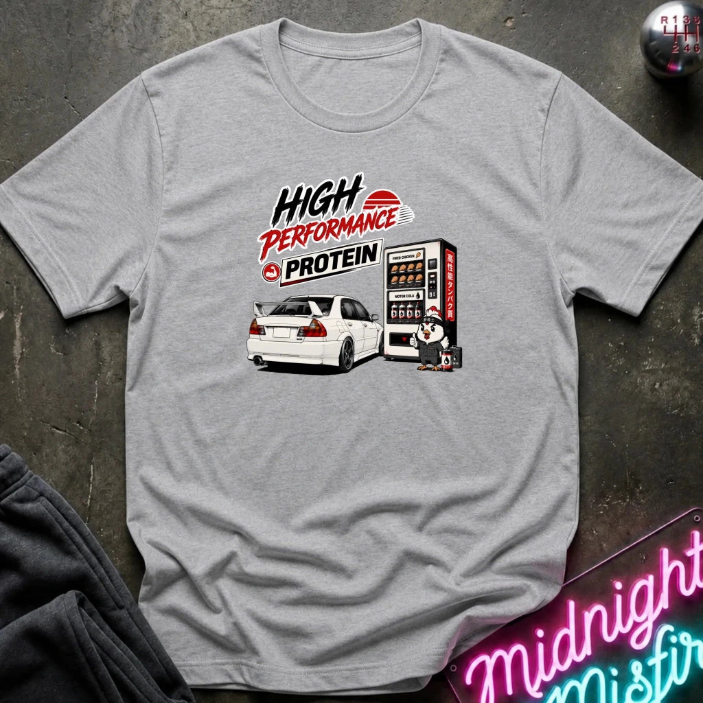A heather gray short-sleeve t-shirt with a graphic print of a white sports car next to a vending machine and the text 'HIGH PERFORMANCE PROTEIN'. The shirt is laid flat on a dark textured surface with a neon sign and dark pants partially visible.