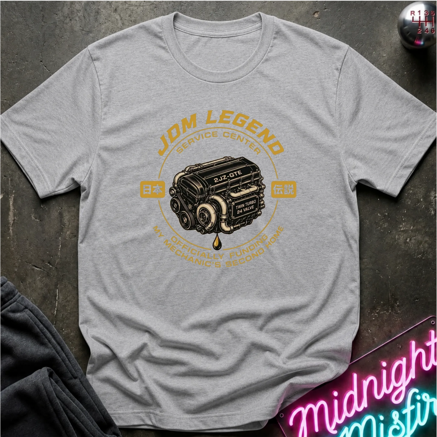 A light gray heather crewneck t-shirt featuring a vintage-style graphic of a JDM engine with the text 'JDM LEGEND' and 'SERVICE CENTER' in yellow and black, laid flat on a dark textured surface.