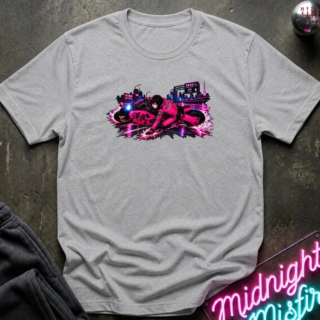 A heather gray short-sleeve t-shirt with a vibrant pink, purple, and blue graphic of a motorcycle and a city skyline on the chest, laid flat on a dark textured surface with a neon sign and sweatpants nearby.