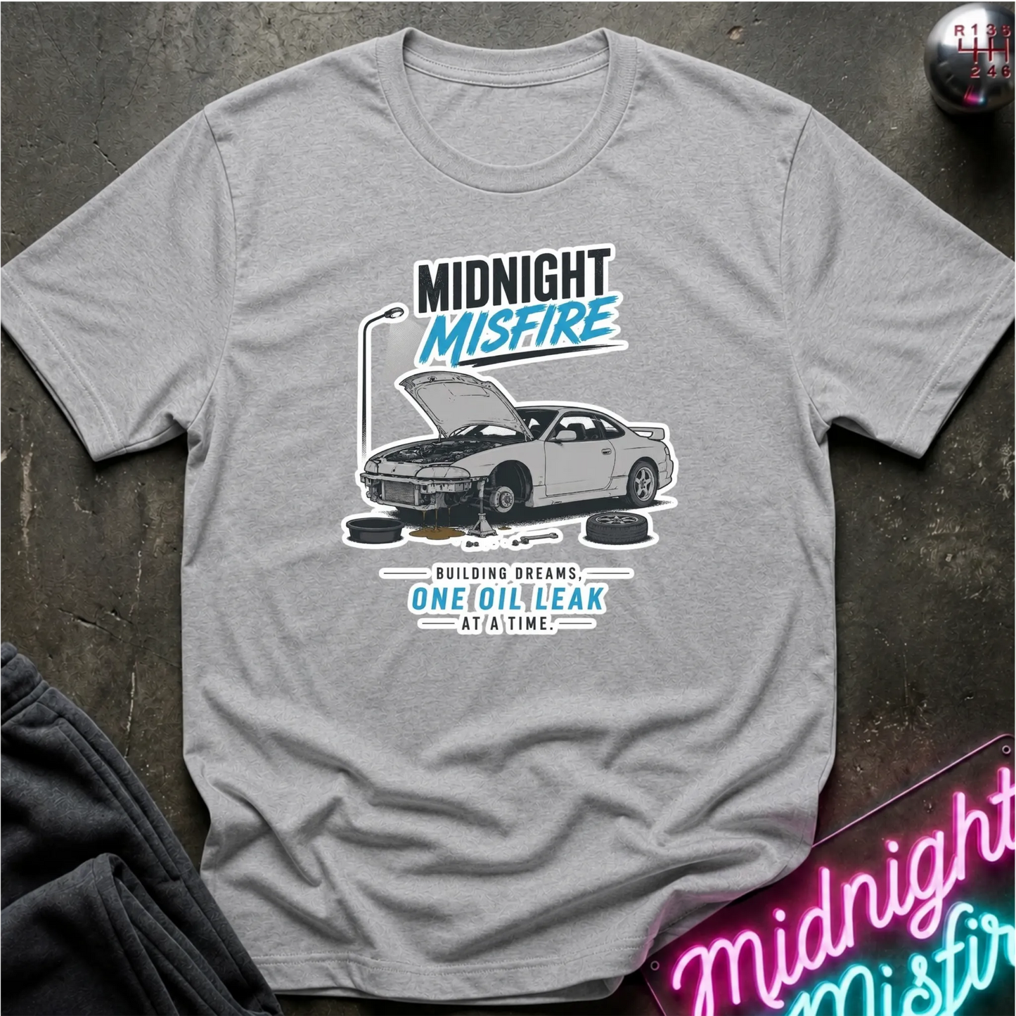 A light gray crewneck t-shirt with a 'Midnight Misfire' car graphic, laid flat on a dark textured surface with a neon sign and dark pants in the background.