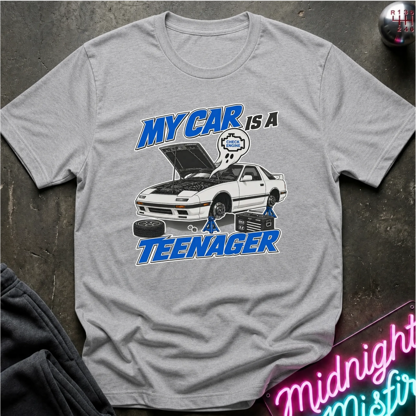 A heather gray short-sleeve graphic T-shirt laid flat on a dark textured surface. The shirt features a white car with its hood open, blue and white text reading 'MY CAR IS A TEENAGER,'.