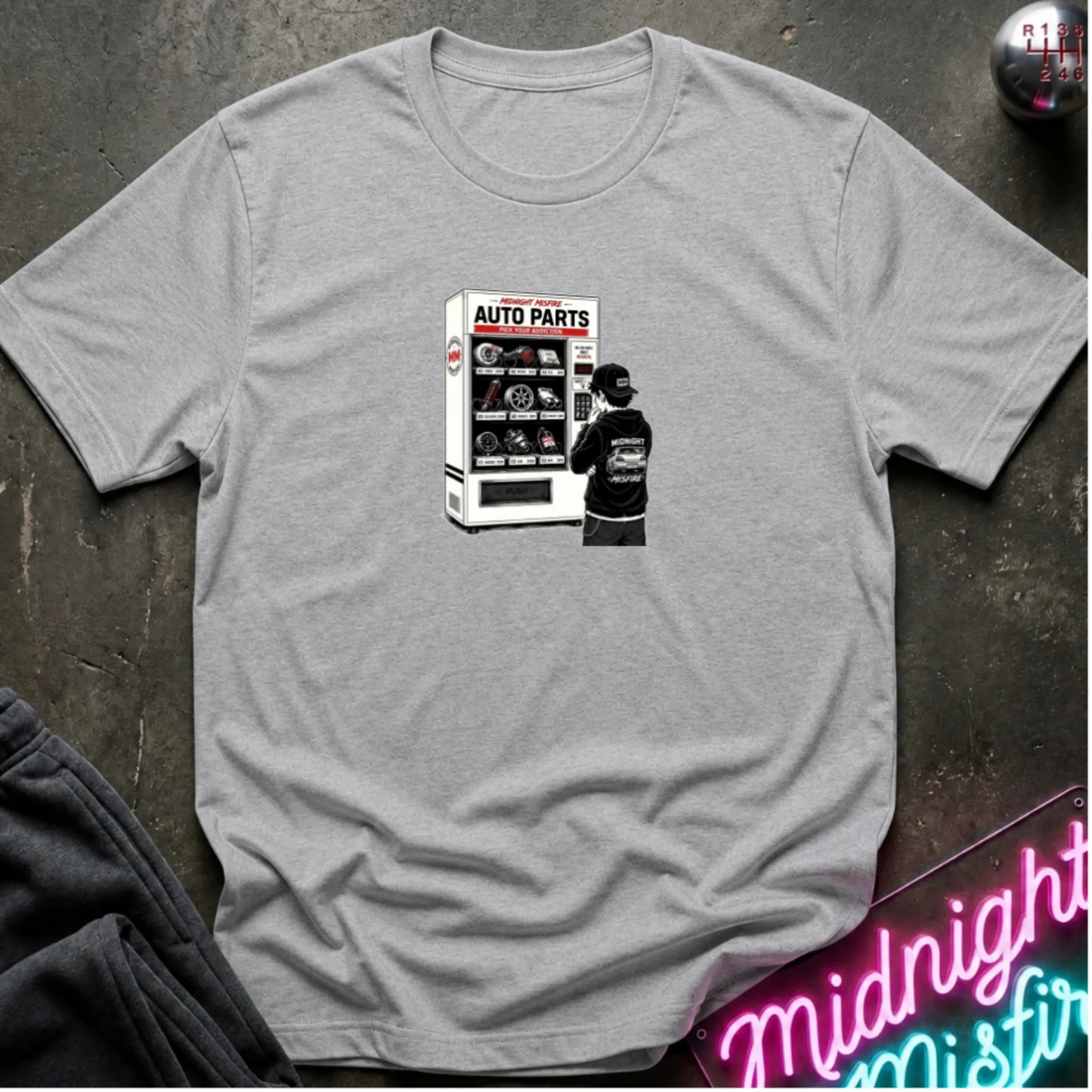 a heather gray short-sleeve t-shirt laid flat on a dark textured surface, featuring a graphic print of a vending machine and owner deciding.