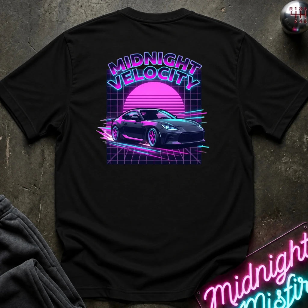 A black short-sleeve t-shirt laid flat on a dark textured surface, showing a large retro 80s synthwave graphic on the back. The graphic features the text 'MIDNIGHT VELOCITY' in pink and blue neon-style lettering above a pink sun and a dark sports car on a grid.