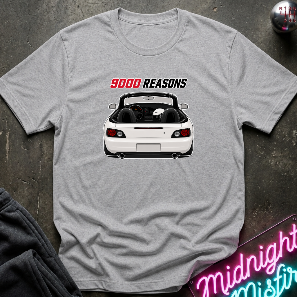 A light gray heather crewneck t-shirt featuring a graphic of a white convertible sports car with the text '9000 REASONS' above it. The shirt is displayed flat on a dark textured surface with a neon sign and dark pants partially visible in the corners.
