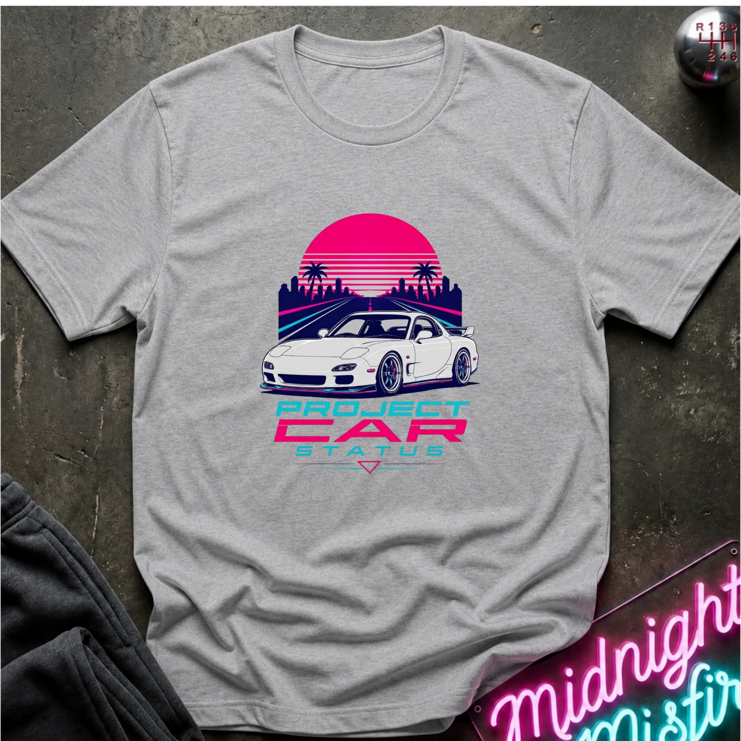 A light gray short-sleeve graphic T-shirt laid flat on a dark textured surface. The shirt features a retro-style print of a white sports car in front of a pink and purple sunset with palm trees, and the text 'PROJECT CAR STATUS' below.