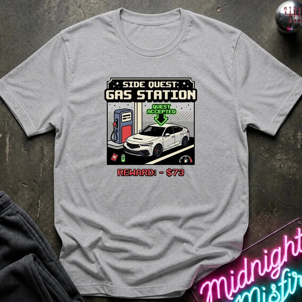 A light gray crewneck t-shirt featuring a retro video game style graphic of a white sports car at a gas station with the text 'SIDE QUEST: GAS STATION' and 'REWARD: $73'. The shirt is displayed flat on a dark textured surface with a neon sign and dark pants partially visible.