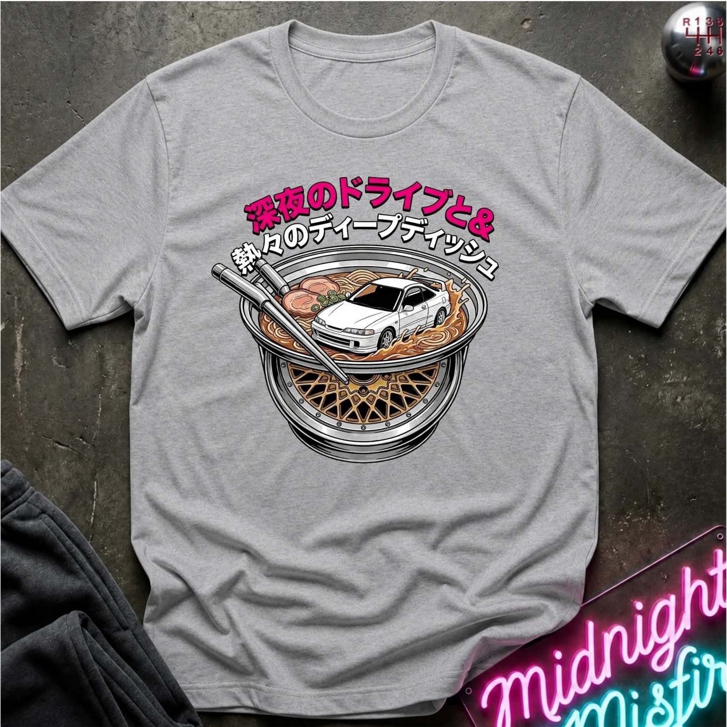 A light gray short-sleeve t-shirt with a graphic print of a white sports car inside a ramen bowl with Japanese text, laid flat on a dark textured surface.
