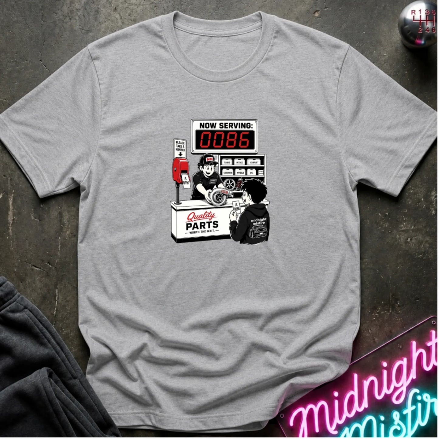 A heather gray  short-sleeve t-shirt with a retro-style graphic of a garage attendant at a 'Quality Parts' counter, displayed flat on a dark textured surface with a neon sign and sweatpants partially visible.