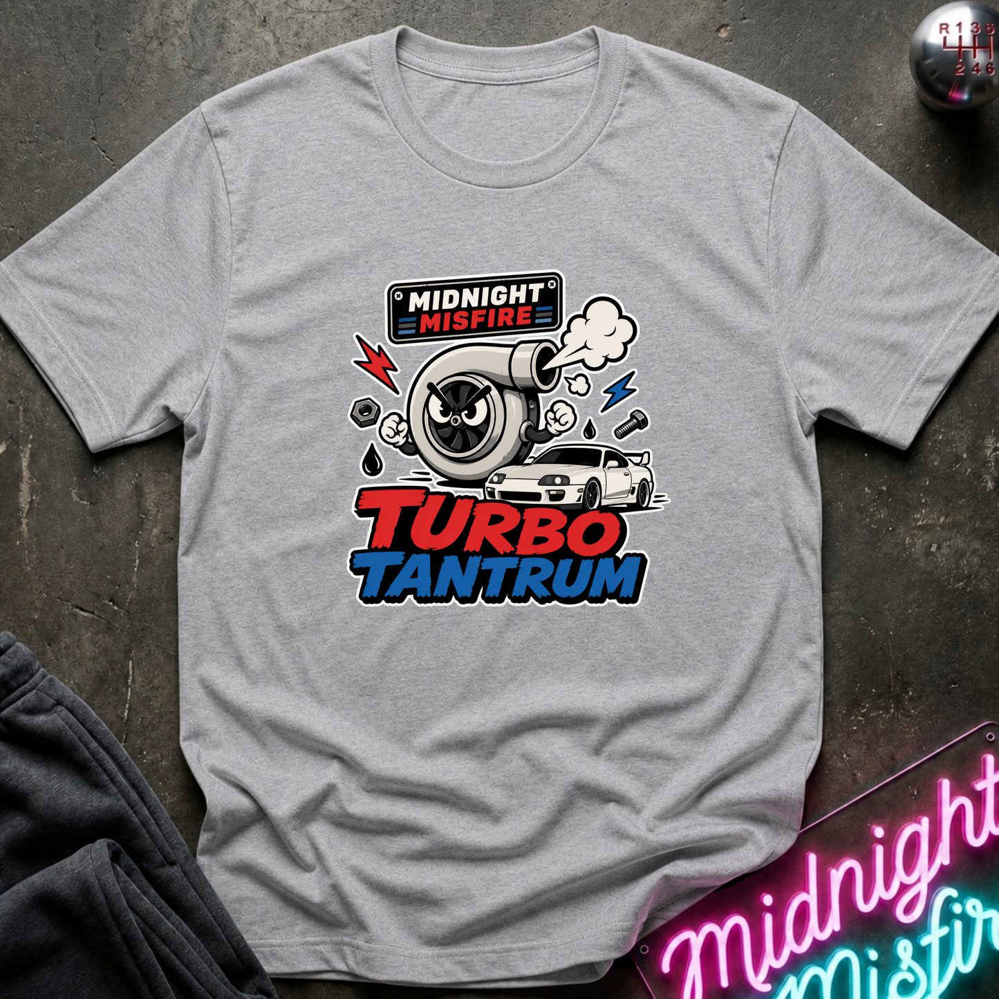 A light gray crewneck t-shirt with a 'Midnight Misfire Turbo Tantrum' graphic on the chest, laid flat on a dark textured surface with a neon sign in the corner.