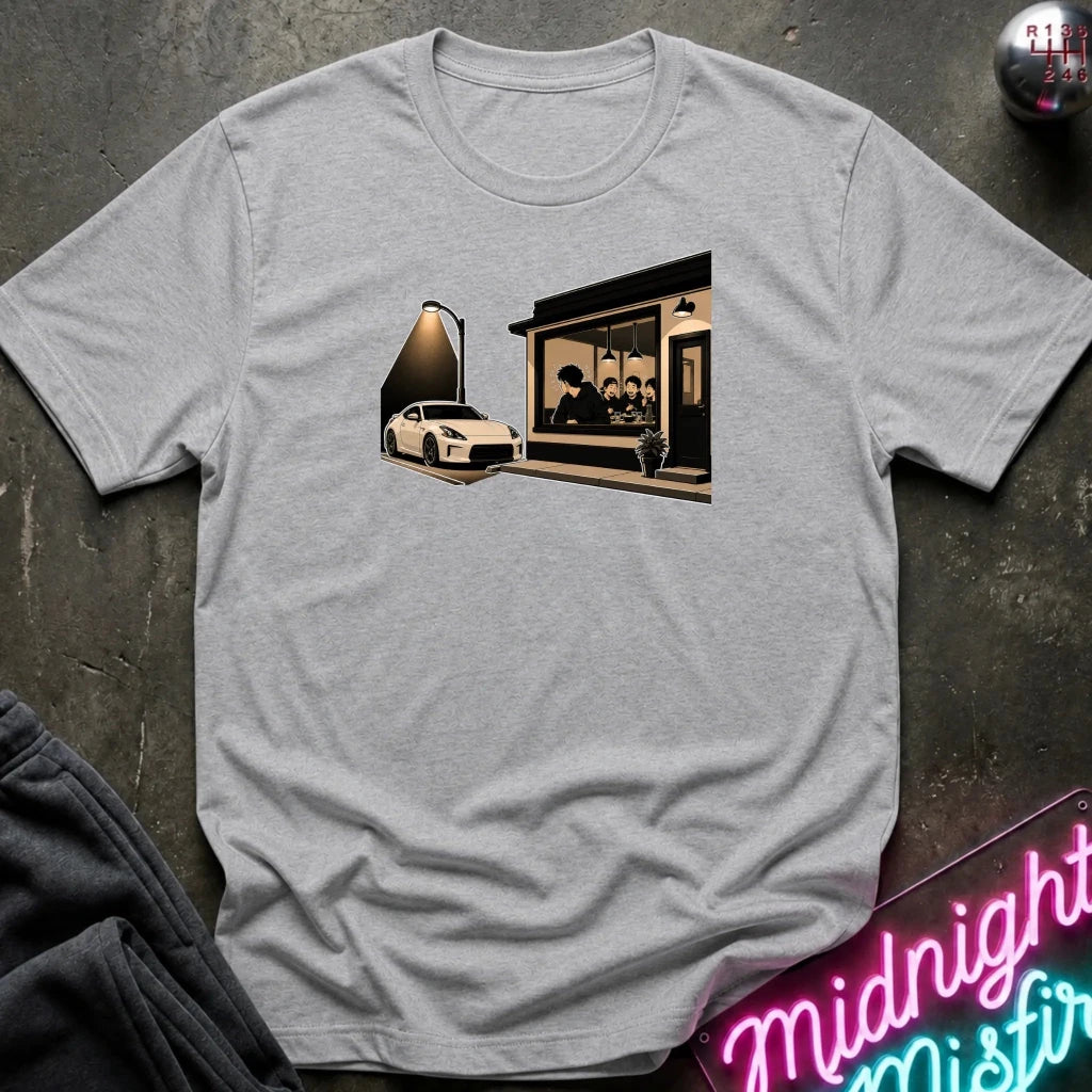 A light heather gray short-sleeve t-shirt laid flat on a dark textured surface. The shirt features a graphic print of a white sports car parked in front of a cafe with a neon sign glowing in the window.