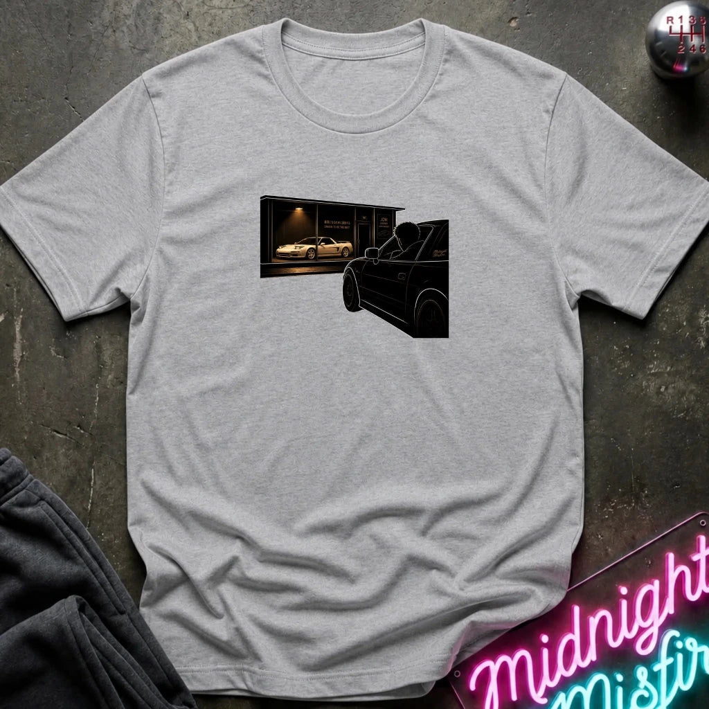 A light heather gray crewneck t-shirt laid flat on a dark textured surface. The shirt features a rectangular graphic print on the chest showing a black convertible car in front of a garage with a white sports car inside.