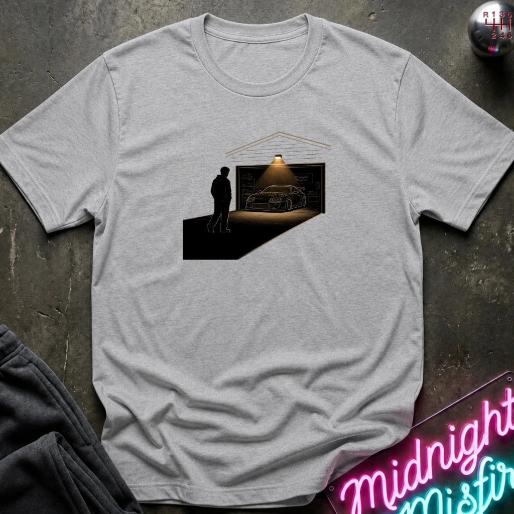 A light heather gray crewneck t-shirt laid flat on a dark textured surface. The shirt features a central graphic print of a silhouette of a man looking at a car inside a garage under a spotlight.