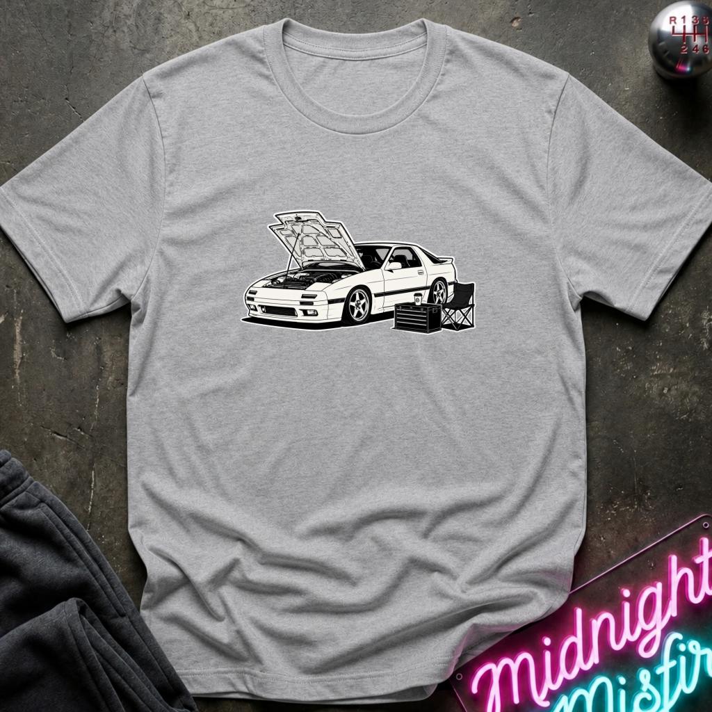 A light gray heather crewneck t-shirt featuring a black and white graphic of a sports car with its hood open, laid flat on a dark textured surface with a neon sign and dark pants partially visible.