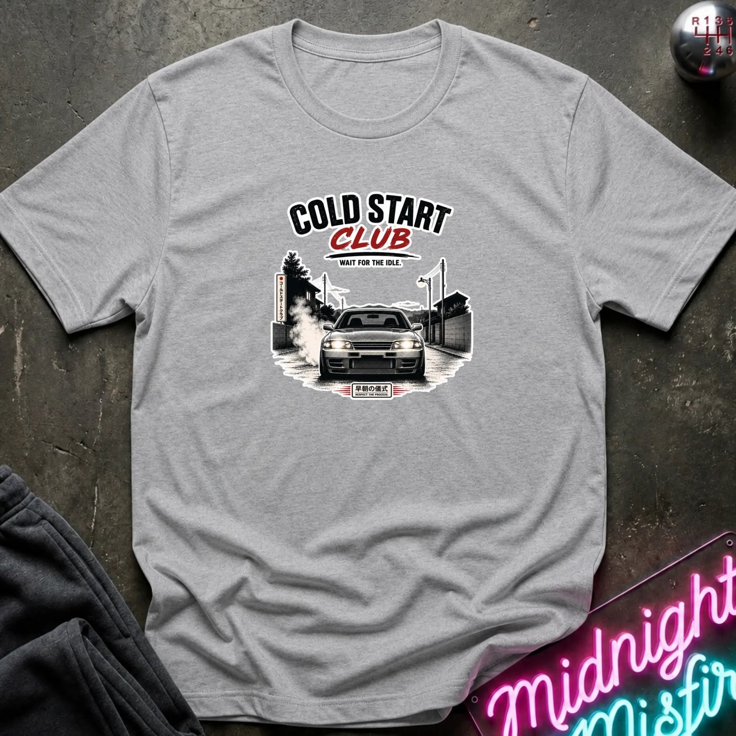 A light gray crewneck t-shirt laid flat on a dark textured surface, featuring a graphic print of a car with the text 'COLD START CLUB''.