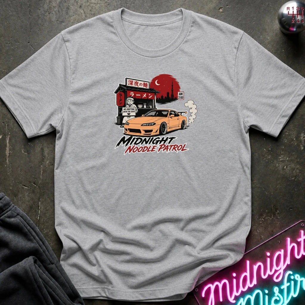 A light gray heather crewneck t-shirt featuring a graphic print of a yellow Nissan Silvia S15 drift car in front of a red moon and Japanese storefront, with the text 'MIDNIGHT Noodle PATROL'. The shirt is laid flat on a dark textured surface with a neon sign and dark pants partially visible.