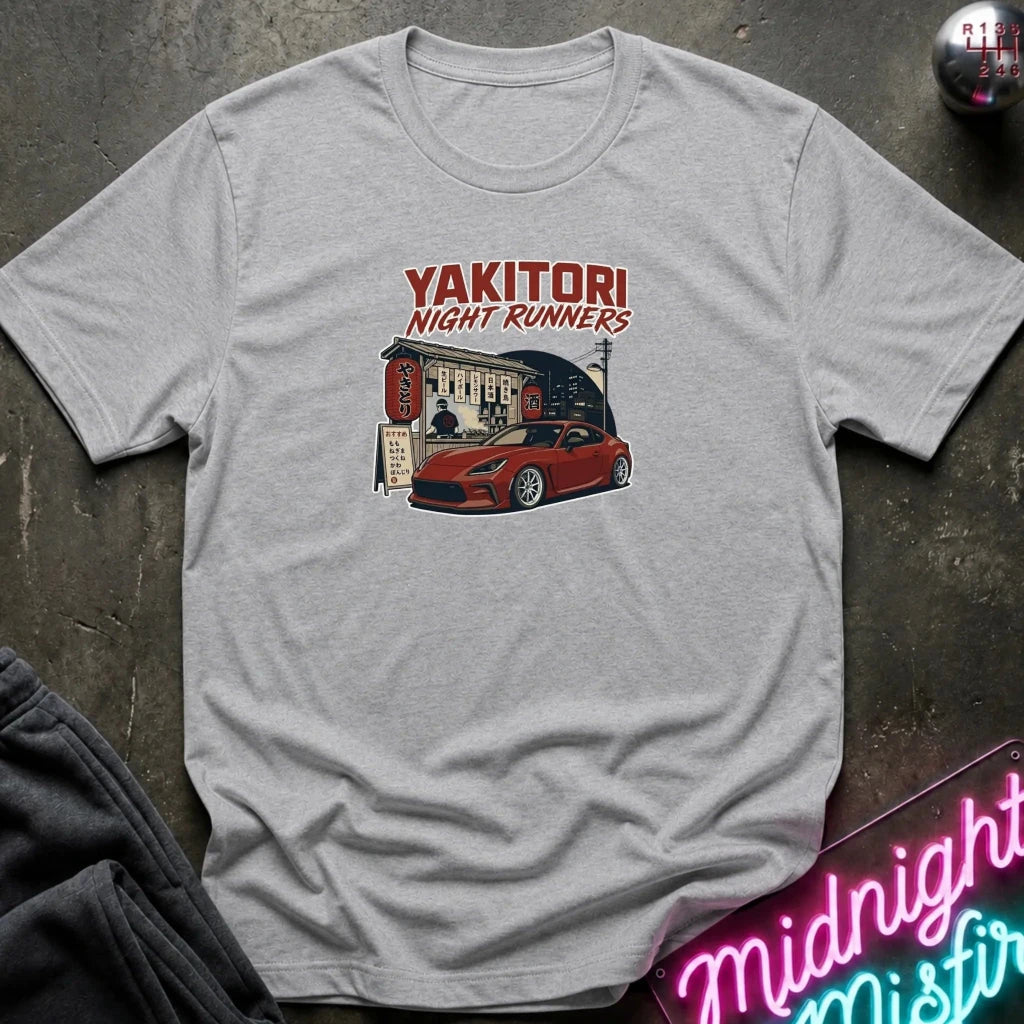 A light gray heather crewneck t-shirt featuring a graphic print of a red sports car parked outside a Japanese restaurant with the text 'YAKITORI NIGHT RUNNERS' above it. The shirt is displayed flat on a dark textured surface with a neon sign and dark pants partially visible in the corners.