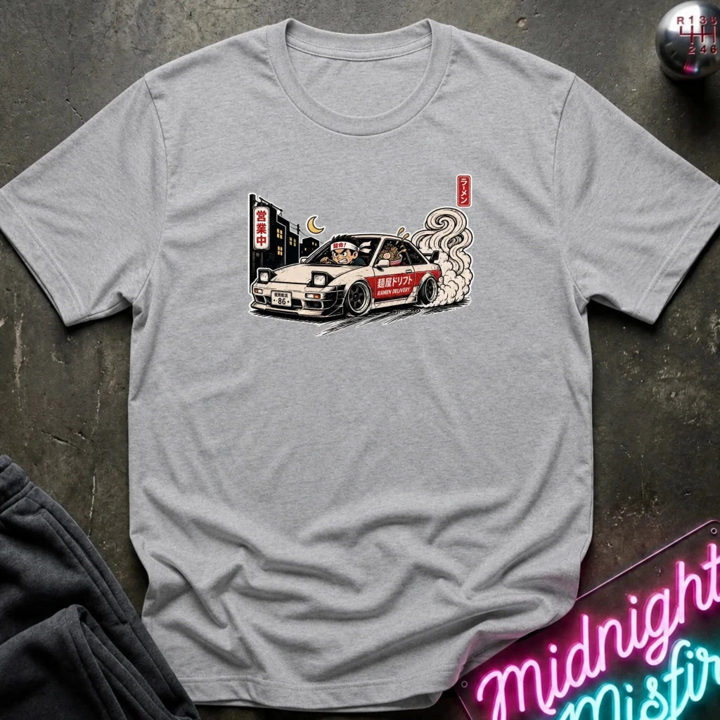 A light gray crewneck t-shirt featuring a graphic print of a white Nissan Skyline R32 race car with a cat driver, styled flat on a dark textured surface with a neon sign and dark sweatpants nearby.