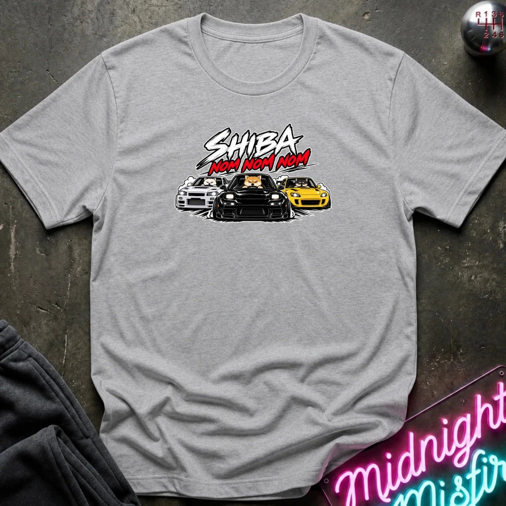 A light gray heather short-sleeve graphic t-shirt laid flat on a dark textured surface. The graphic features the text 'SHIBA NOM NOM NOM' in white and red, above an illustration of three cars (white, black, and yellow).