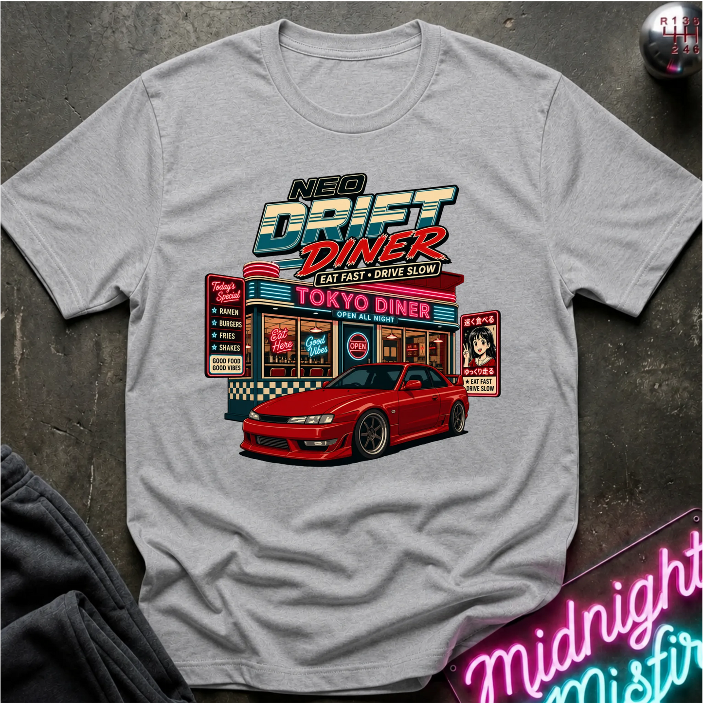 A light gray crewneck t-shirt featuring a colorful graphic print of a retro 'Neo Drift Diner' with a red sports car in front, laid flat on a dark textured surface.