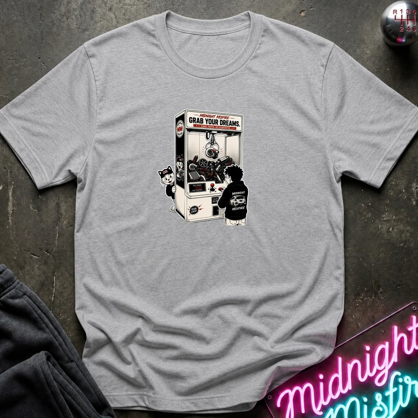 A light gray heather short-sleeve crewneck t-shirt laid flat on a dark textured surface, featuring a central graphic of a person in a black hoodie playing a claw machine. 