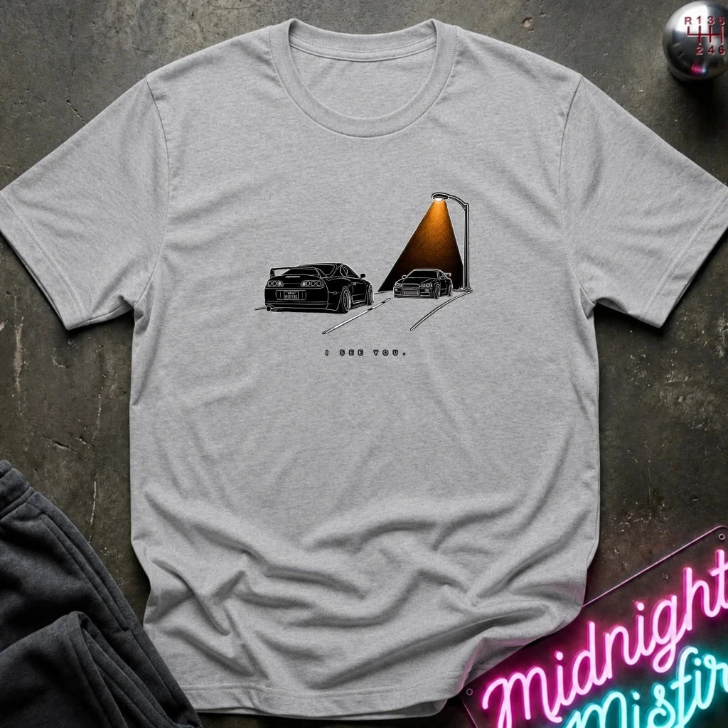 A light gray heather short-sleeve t-shirt laid flat on a dark textured surface, featuring a graphic print of two black sports cars passing each other.