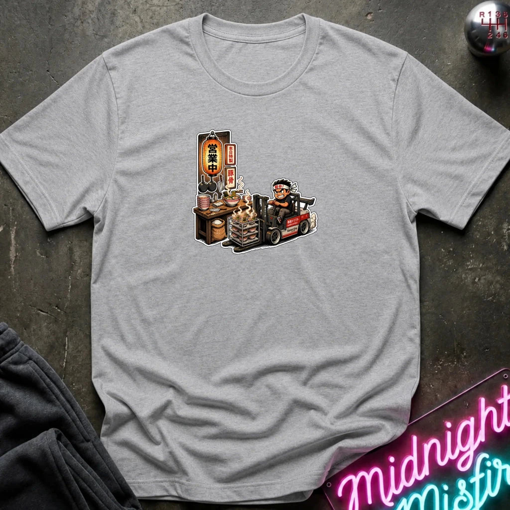 A light heather gray crewneck t-shirt featuring a cartoon graphic of a forklift driver working at a street food stall, laid flat on a dark textured surface with a neon sign and dark pants partially visible.
